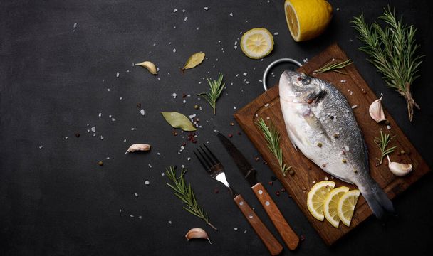 Raw whole dorado fish on wooden kitchen board and spices for cooking, top view on black table - 写真・画像