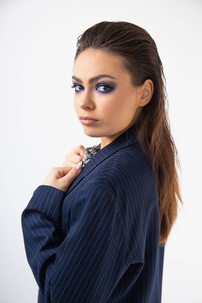 Modern beautiful young woman with loose long hair in blue striped jacket looks half-turned into camera. Bright blue makeup. Professional makeup artist. Close-up. Studio, white background. Fashion. - Foto, afbeelding