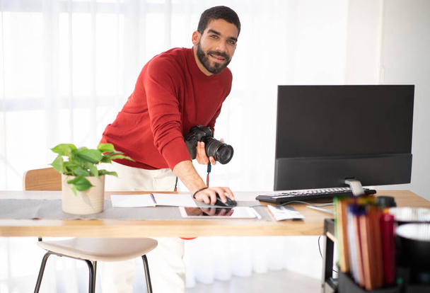 Photographe professionnel joyeux beau barbu hispanique jeune homme qui travaille sur la séance photo au studio, tenant un appareil photo dslr, en utilisant un ordinateur PC, souriant à la caméra, copier l'espace - Photo, image