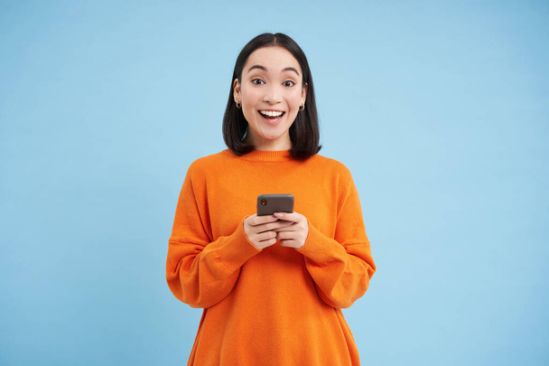 Glückliche Asiatin mit Smartphone, tolles Angebot online auf Handy gefunden, begeistert über Rabatte in App, steht vor blauem Hintergrund. - Foto, Bild