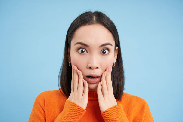 Close up portrait of asian girl with shocked face, looks at smth unbelievable, startled by news, stands over blue background. - Photo, Image