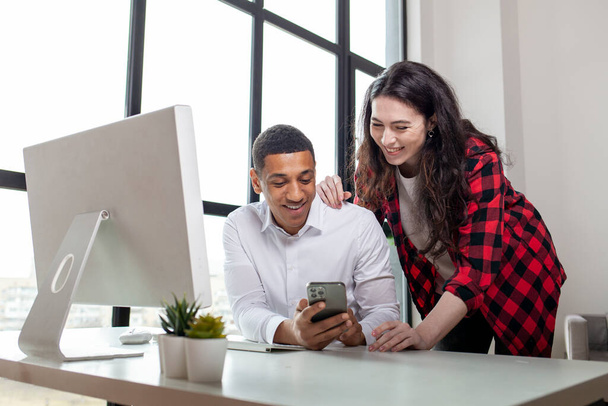 african american man in business clothes and glasses sits at computer with his wife and uses smartphone, young european woman stands and supports her husband, multiracial couple looks at the phone - Photo, Image