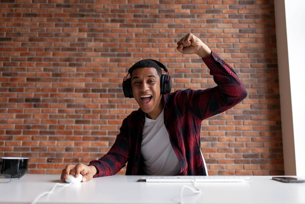 joyful guy african american gamer plays computer game and rejoices in victory, male winner sits at computer table and celebrates success - 写真・画像
