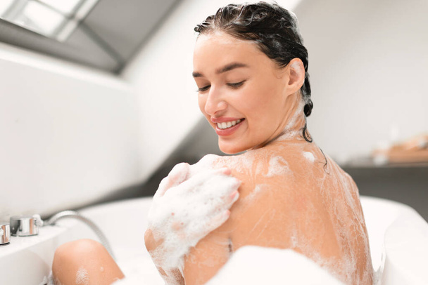 Happy Woman Washing Her Body Touching Smooth Skin With Applied Shower Gel Taking Bath Indoors. Lady Rubbing Shoulder Covered With Foam Bathing In Bathroom. Selective Focus - Foto, Imagem
