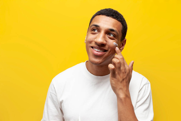 young african american guy in white t-shirt applies cream on his face on yellow isolated background, man uses facial cosmetics and takes care of his skin - Фото, изображение