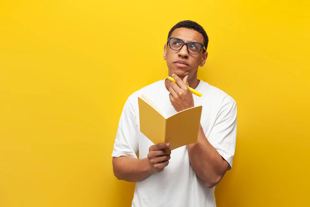 pensive guy african american student in white t-shirt and glasses writes in notebook and plans on yellow isolated background, man dreams and imagines - Photo, Image