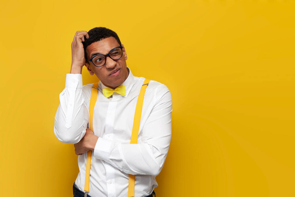 young pensive guy african american in white shirt with suspenders and bow tie scratches his head on yellow isolated background, nerd man in glasses remembers - Foto, Imagen