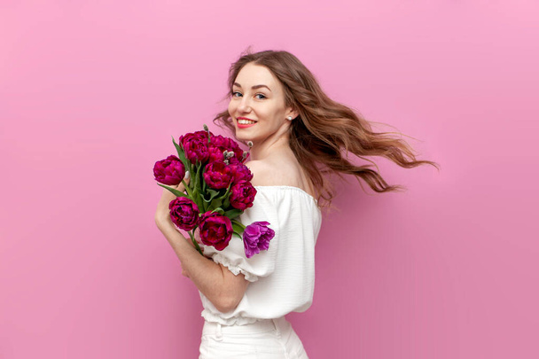 young attractive girl in white clothes holds bouquet of pink tulips and smiles, woman on holiday with flowers on pink isolated background, concept of spring and March 8 - Photo, Image