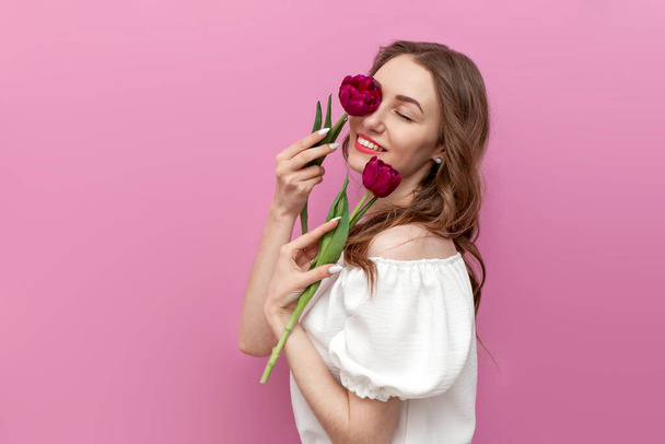 молодая нежная девушка прикасается к коже лица двумя розовыми тюльпанами и улыбается на розовом изолированном фоне, женщина с закрытыми глазами в белой одежде позирует с цветами - Фото, изображение