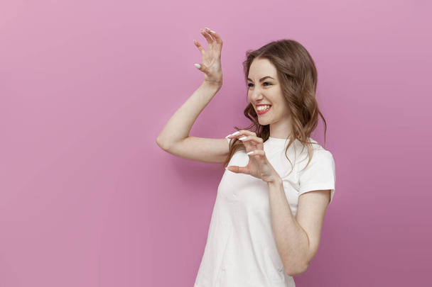 young cute woman in white t-shirt shows her claws with her fingers and imitates tiger, the girl growls and bares her teeth on pink isolated background - Φωτογραφία, εικόνα