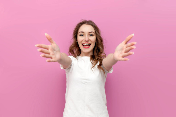 young cute woman in white t-shirt with outstretched arms wants hug, the girl hugs and holds her hands in front of her on pink isolated background - Photo, image
