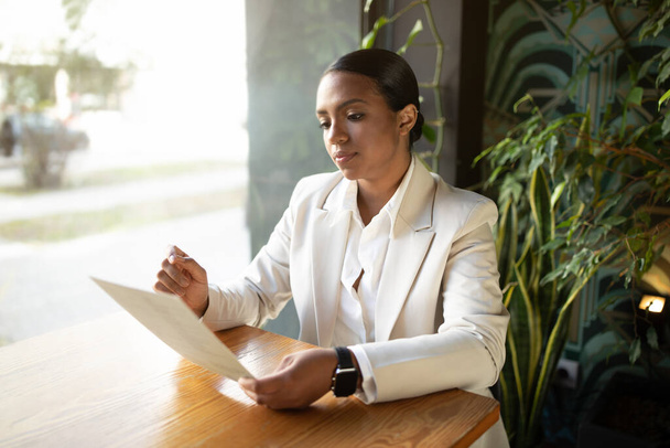 Serious concentrated millennial african american businesswoman in white suit reading document in cafe office with green plants interior. Startup, modern business, work and lifestyle - Foto, Imagen