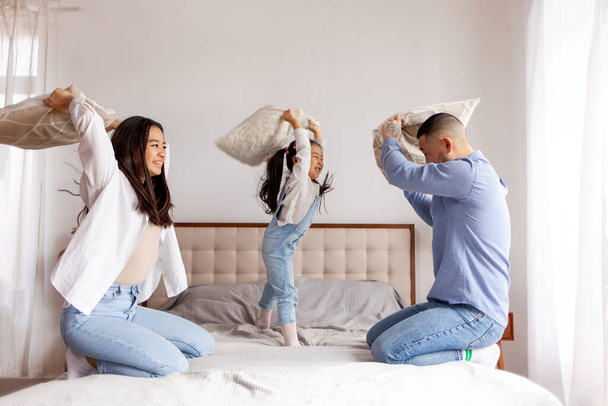 Kissenschlacht, kleines asiatisches Mädchen spielt mit ihren Eltern zu Hause auf dem Bett, koreanische Eltern verbringen Zeit mit ihrer Tochter und freuen sich, Mama und Papa haben Spaß mit dem Kind - Foto, Bild