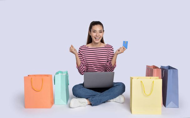 Emotionele stijlvolle vrolijke glimlachende jonge brunette vrouw zittend op de vloer tussen kleurrijke winkeltassen, met behulp van laptop, met bankkaart en gebaren, grijze studio achtergrond. E-commerce, retail concept - Foto, afbeelding