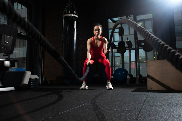 athletic woman in red sportswear trains with ropes in black gym, girl does fitness in fitness room, attractive woman in fitness club on crossfit workout - Photo, image