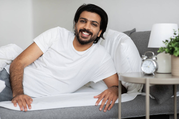Joyeux jeune homme du Moyen-Orient en t-shirt blanc se trouve sur le lit, profiter du temps libre et bonne matinée dans la chambre à coucher intérieur avec réveil, espace libre. Week-end seul et vitalité à la maison - Photo, image