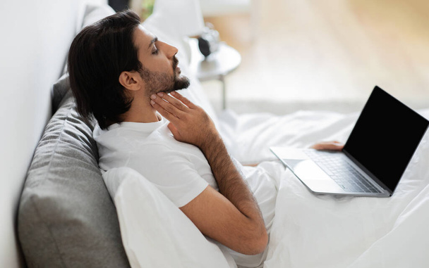 El joven árabe infeliz en camiseta blanca se sienta en la cama, comprueba las amígdalas, tiene videollamada en la computadora portátil con pantalla en blanco, sufre de gripe y frío en el interior del dormitorio. Consulta en línea, tratada en casa - Foto, imagen
