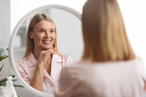Концепция Skincare. Привлекательная женщина средних лет, смотрящая на зеркало в ванной комнате, красивая зрелая женщина, трогающая лицо и улыбающаяся своему отражению, наслаждающаяся своей безупречной кожей, избирательный фокус - Фото, изображение