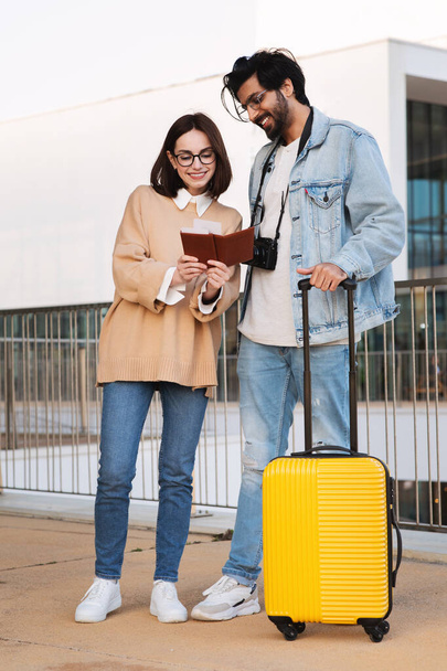 Glad millennial multiethnic family tourists with yellow suitcase look at passport, enjoy trip in airport in city, vertical, full length. Relationship, travel, vacation together and people emotions - Photo, Image