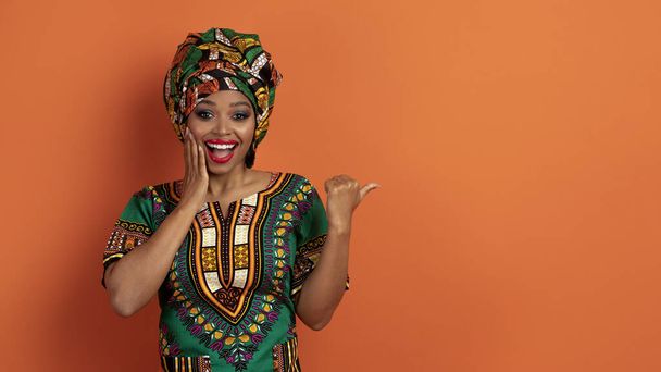 Niesamowite wesoły piękne młoda czarna kobieta w kolorowe tradycyjne afrykańskie ubrania pokazujące przestrzeń do kopiowania reklamy i dotykania jej twarzy, izolowane na pomarańczowym tle studio, panorama - Zdjęcie, obraz