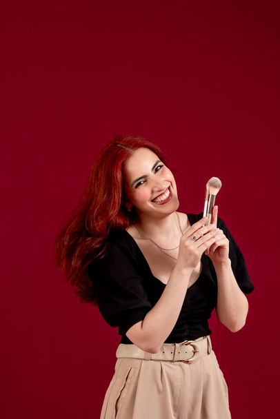 Woman looking at camera and smiling while holding makeup brushes standing against an isolated background. Beauty and cosmetic concept. Vertical picture. - Foto, immagini