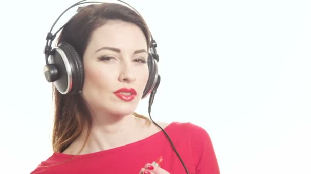 Mooi meisje in de rode luisteren naar de muziek in grote hoofdtelefoon en dansen met plezier toepassing van rode lippenstift geïsoleerd op een witte achtergrond close-up shot - Video