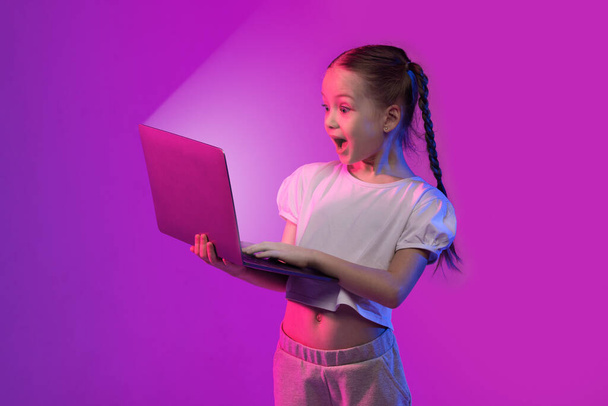 Amazed excited pretty stylish school-aged kid girl looking at shining laptop screen and screaming, checking newest breathtaking video game, colorful background with neon light effect - Photo, image