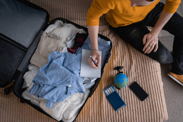 Making a check list of things to pack for travel. Top view of man writing paper take note and packing suitcase to vacation, prepare clothes into luggage. Travel vacation concept - Photo, image