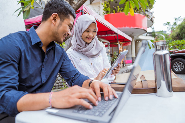 Asian female worker in hijab and male worker using digital tablet and laptop sitting in outdoor background - Fotoğraf, Görsel