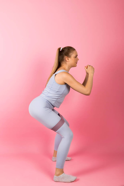 Young healthy Girl in gray sportswear is doing stretches and warm-up exercises with a rubber band resistance. Isolated on pink background. - 写真・画像