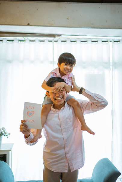 young asian dad shoulders piggybacks his son showing a paper love drawing during playing at home - Fotoğraf, Görsel