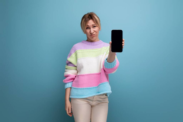 angenehme blonde junge Frau im gestreiften Pullover demonstriert eine Smartphone-Attrappe auf blauem Hintergrund. - Foto, Bild
