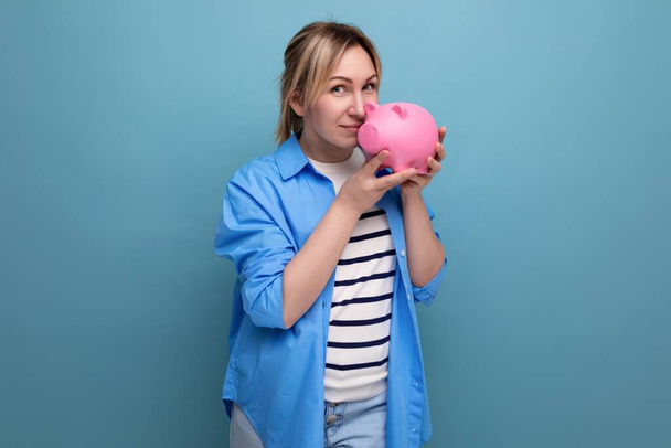 blond usměvavá dívka v ležérní oblečení drží prasátko banky na modrém pozadí s kopírovacím prostorem. - Fotografie, Obrázek