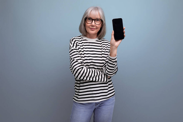 gepflegte schlanke Frau mittleren Alters in den 60ern hält ein Smartphone mit einer Attrappe auf hellem Hintergrund mit Kopierraum. - Foto, Bild
