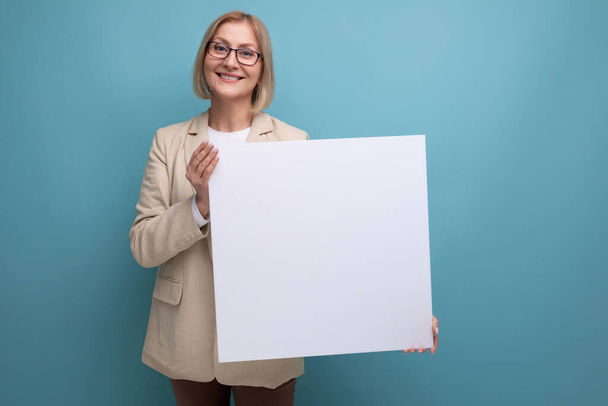 усміхнена зріла жінка в куртці демонструє порожній макет паперу на студійному фоні з копіювальним простором. - Фото, зображення
