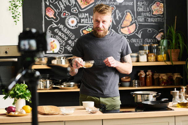Σύγχρονη blogger αρσενικό φαγητό με γενειάδα στο πρόσωπο κρατώντας γυάλινο μπολ με τηγανητά σουσάμι σπόρους μιλώντας για το πιάτο συνταγή στην κάμερα - Φωτογραφία, εικόνα