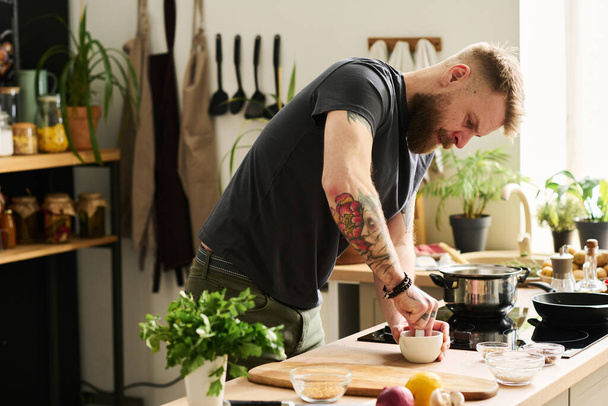Knappe jonge blanke man met tatoeages op armen staande in keuken verpletterende specerijen met mortel en stamper - Foto, afbeelding