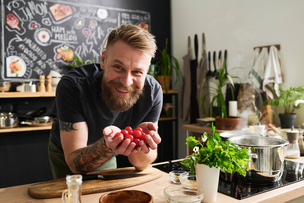 Yüzünde sakalı ve kollarında dövmeleri olan neşeli beyaz bir adam ellerinde vişneli domateslerle kameraya gülümsüyor. - Fotoğraf, Görsel