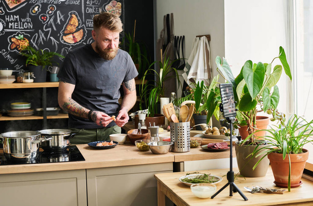 Красивый бородатый белый мужчина, стоящий на чердаке кухни и готовящий ужин на камере, очищающий креветки. - Фото, изображение
