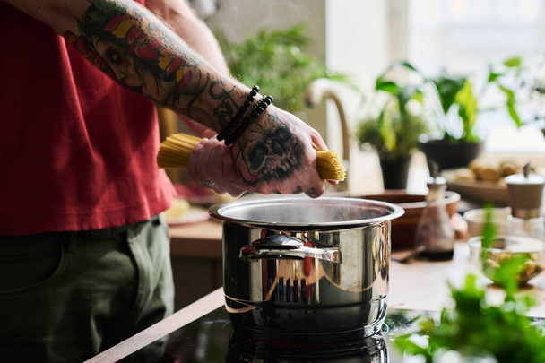 Hands of unrecognizable tattooed man putting spaghetti into stainless steel pot with boiling water - Photo, Image
