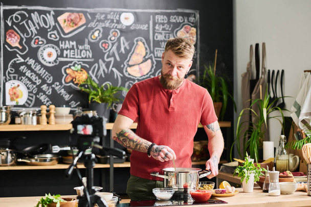 Όμορφος άνδρας με τατουάζ Καυκάσιος με γενειάδα στο πρόσωπο στέκεται στη σοφίτα κουζίνα μαγειρεύοντας ζυμαρικά και μιλώντας στην κάμερα για φαγητό blog - Φωτογραφία, εικόνα