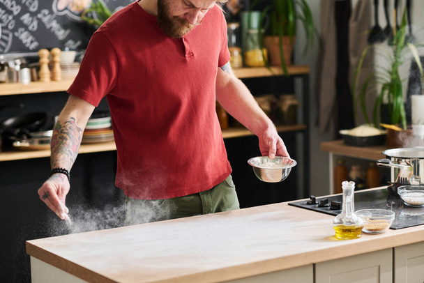 Modern profesyonel şef gündelik kıyafetlerle mutfak masasının tozunu alırken hamur yapmak için yemek pişiriyor. - Fotoğraf, Görsel
