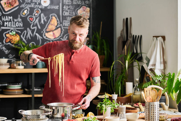Cheerful Caucasian man with beard on face taking noodles for pasta out of hot water looking at camera - Photo, Image