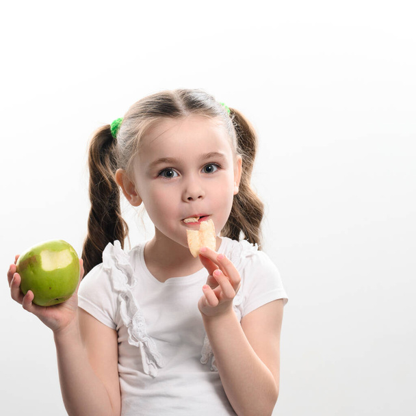 Pieni tyttö pitelee yhtä omenaa ja sipsejä valkoisella taustalla, terveellisten ja epäterveellisten ruokavalintojen käsite lapsen elämässä.. - Valokuva, kuva