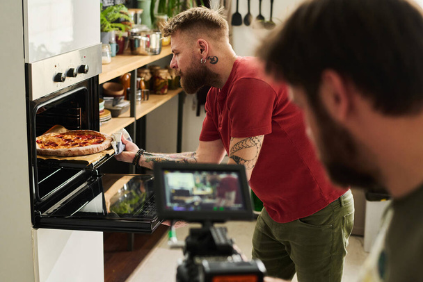 Nierozpoznawalny kamerzysta filmujący współczesnego brodatego białego mężczyznę biorącego pizzę z gorącego piekarnika - Zdjęcie, obraz