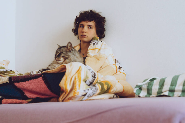 Εφηβικό κορίτσι τυλιγμένο σε κουβέρτα με γάτα, κάθεται στο κρεβάτι, λυπημένο, άρρωστο ή καταθλιπτικό - Φωτογραφία, εικόνα