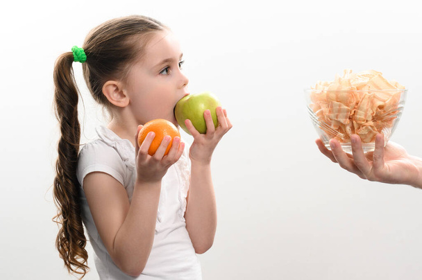 Big bowl of chips and fruit, beautiful little girl eats an apple, child is given a bowl of chips, white background and copy space, healthy and junk food concept. - Foto, imagen
