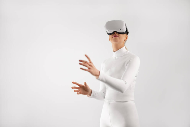 Улыбающаяся женщина, использующая VR гарнитуру, чтобы путешествовать в мире метавселенной виртуальной реальности. Минималистическая концепция с белой одеждой и фоном. - Фото, изображение