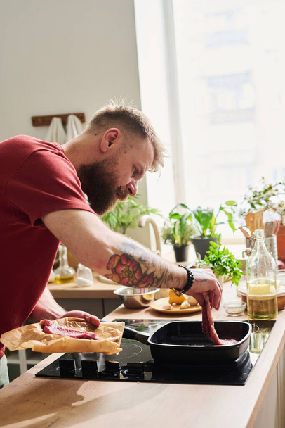 Moderne blanke man met getatoeëerde armen koken rundvlees biefstuk op hete grill pan in loft keuken thuis - Foto, afbeelding