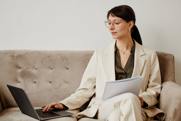 Retrato mínimo de la mujer joven elegante usando el ordenador portátil en el sofá mientras que maneja negocio acertado, espacio de la copia - Foto, imagen
